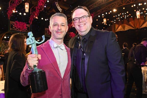 Jeremy Strong, in a fuchsia-colored dinner jacket, holds a statuette at an awards ceremony. Duncan Crabtree-Ireland stands next to him in purple formal wear.