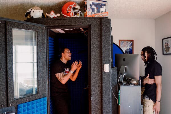 A man in a black T-shirt gestures in a soundproof booth with the door open as a second man in a T-shirt stands outside opposite a video screen.